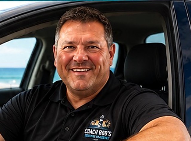 Coach Rod (Mitch Rodrigue) smiling from the driver's seat of his driving-academy vehicle